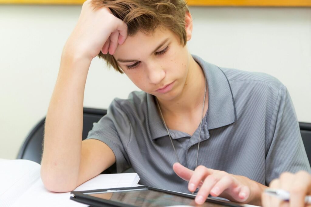 Teenage student with head in hand looking stressed while using a tablet device at a classroom desk