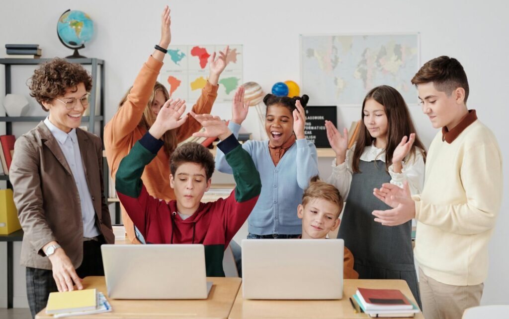 Diverse group of students and teacher celebrating with raised hands around laptops in bright classroom setting
