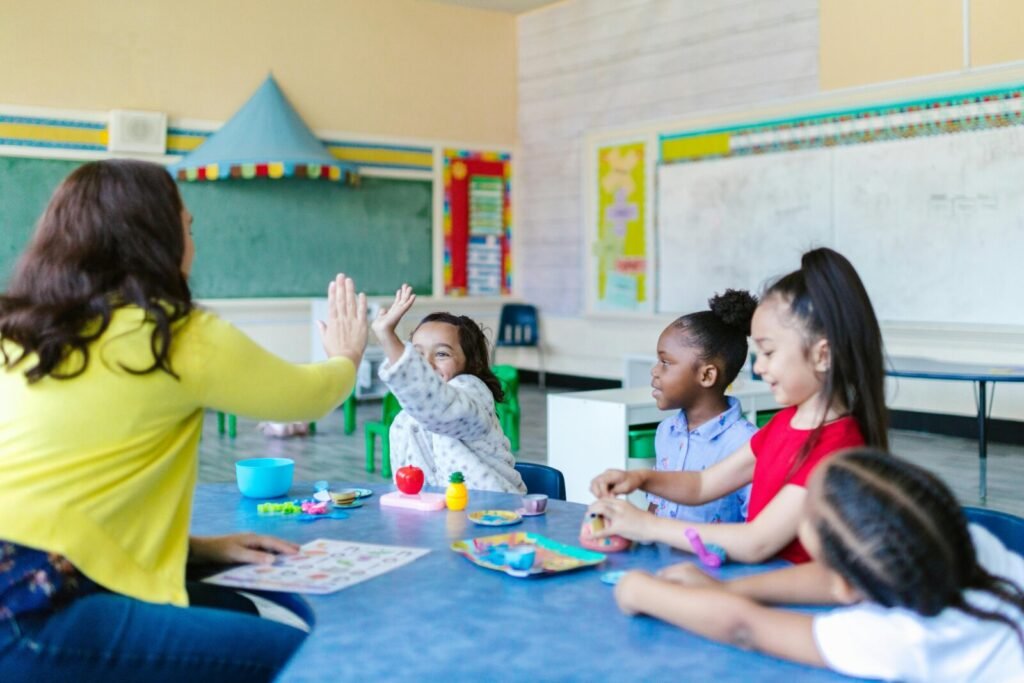Teacher in yellow shirt high-fiving enthusiastic student while other children sit at table in classroom