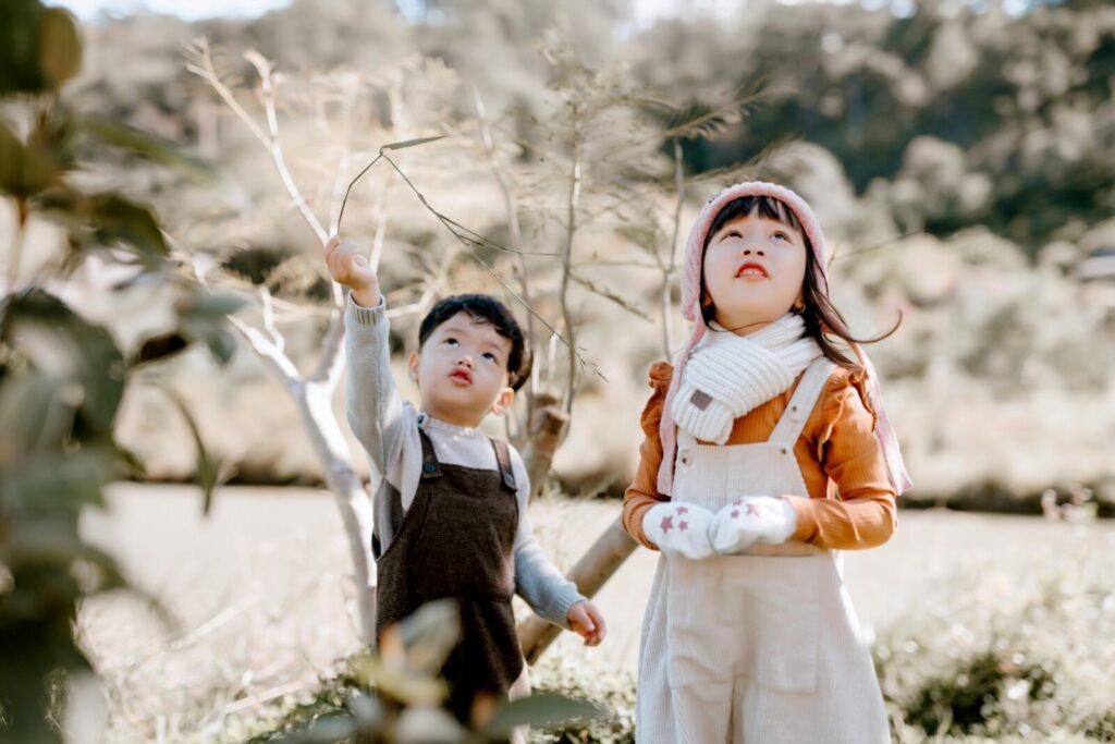 Two children play outdoors among rocks, one throwing sticks upward while another stands nearby