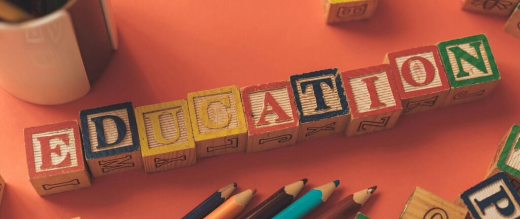 Wooden alphabet blocks spell out "EDUCATION" with colorful pencils scattered nearby on an orange surface.