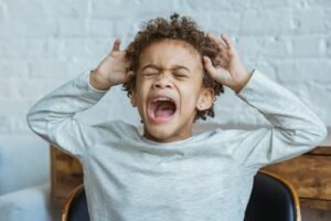 Young child with curly hair screaming with mouth open and hands covering ears