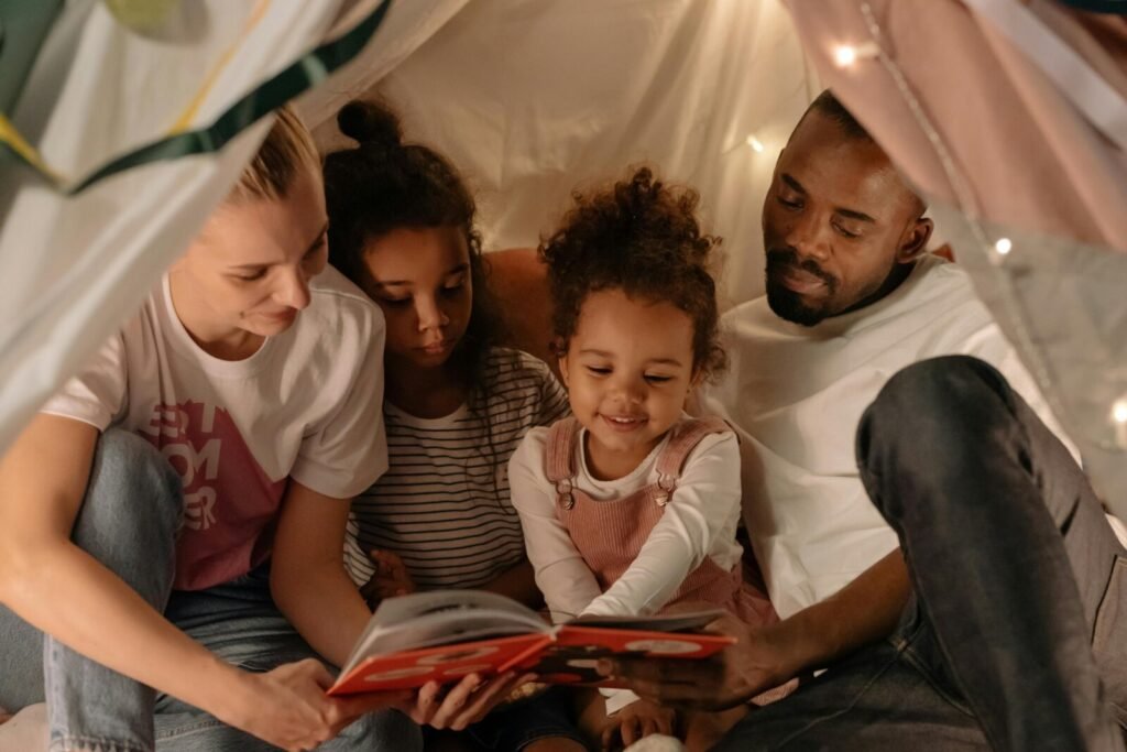 Family of four reading a book together inside a blanket fort illuminated by string lights.