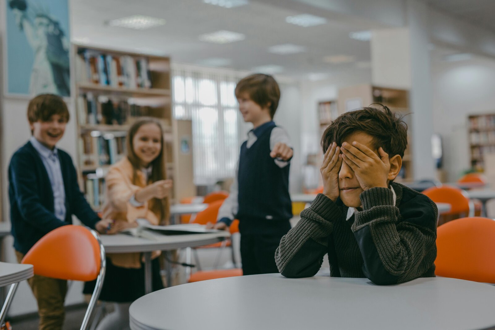 Student sits alone covering face with hands while other students laugh in the background